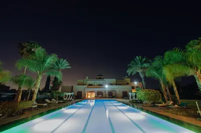 a swimming pool with palm trees in front of a building at Palais les sources de l'atlas, Padel & spa in Marrakech